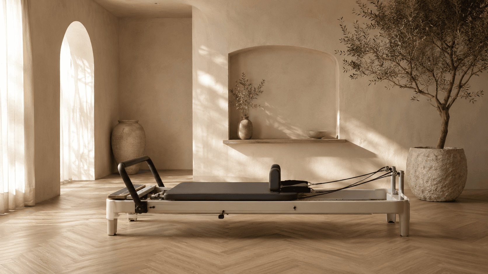 A single wooden reformer in a sunlit arched room, styled with an olive branch and stone vessels.